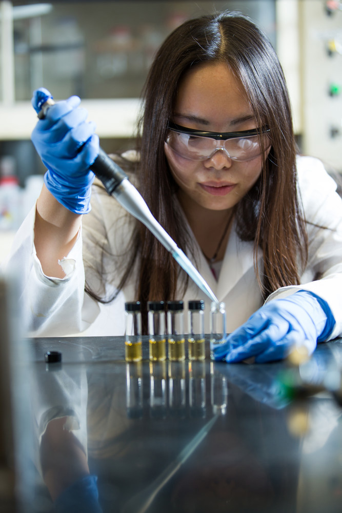 Engineer wearing a lab coat is using a pipette to drop a substance into a small container.